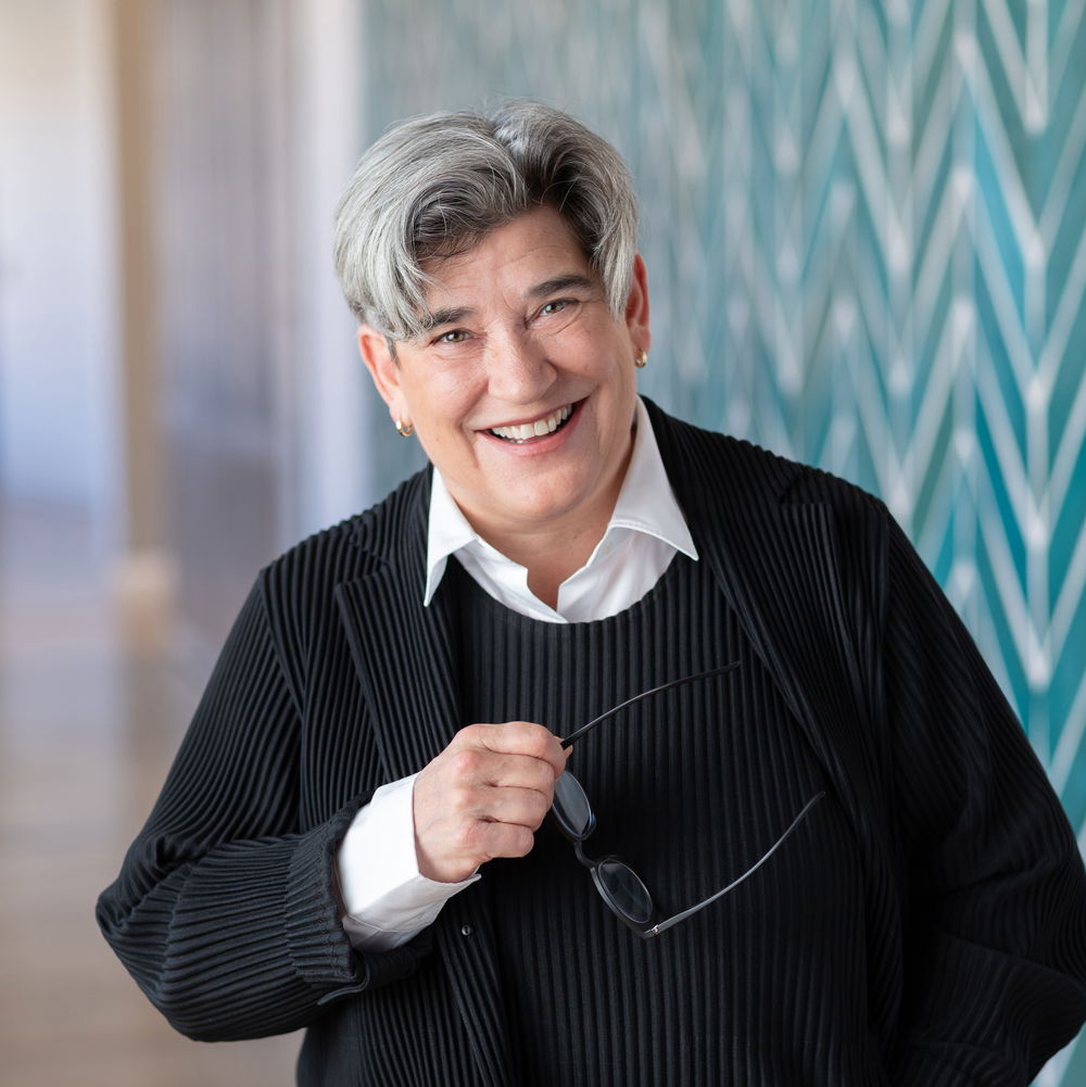 A white woman with short, salt-and-pepper hair smiles widely, holding a pair of glasses in one hand. She stands in front of a patterned wall.