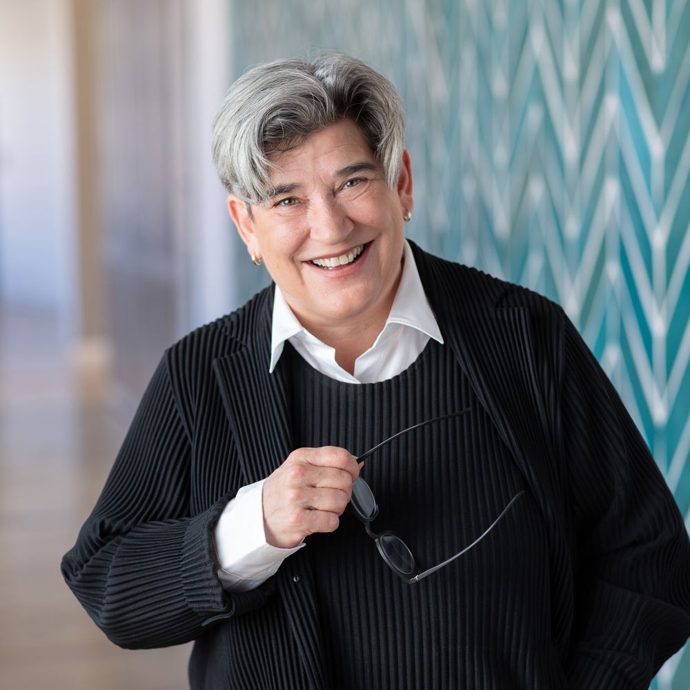 A white woman with short, salt-and-pepper hair smiles widely, holding a pair of glasses in one hand. She stands in front of a patterned wall.