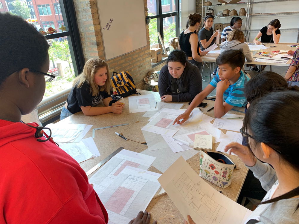 A group of high school students leaning over on a wooden table working collaboratively on a conceptual design. Papers detailing the floorplan are scattered across the table.