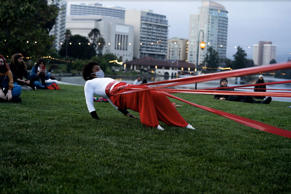 A photograph of an outdoor performance on a waterfront. A performer leans back, supported by taut red ribbons tied at their waist that stretch beyond the frame to the right. The performer wears a white shirt, red pants, and a medical mask.
