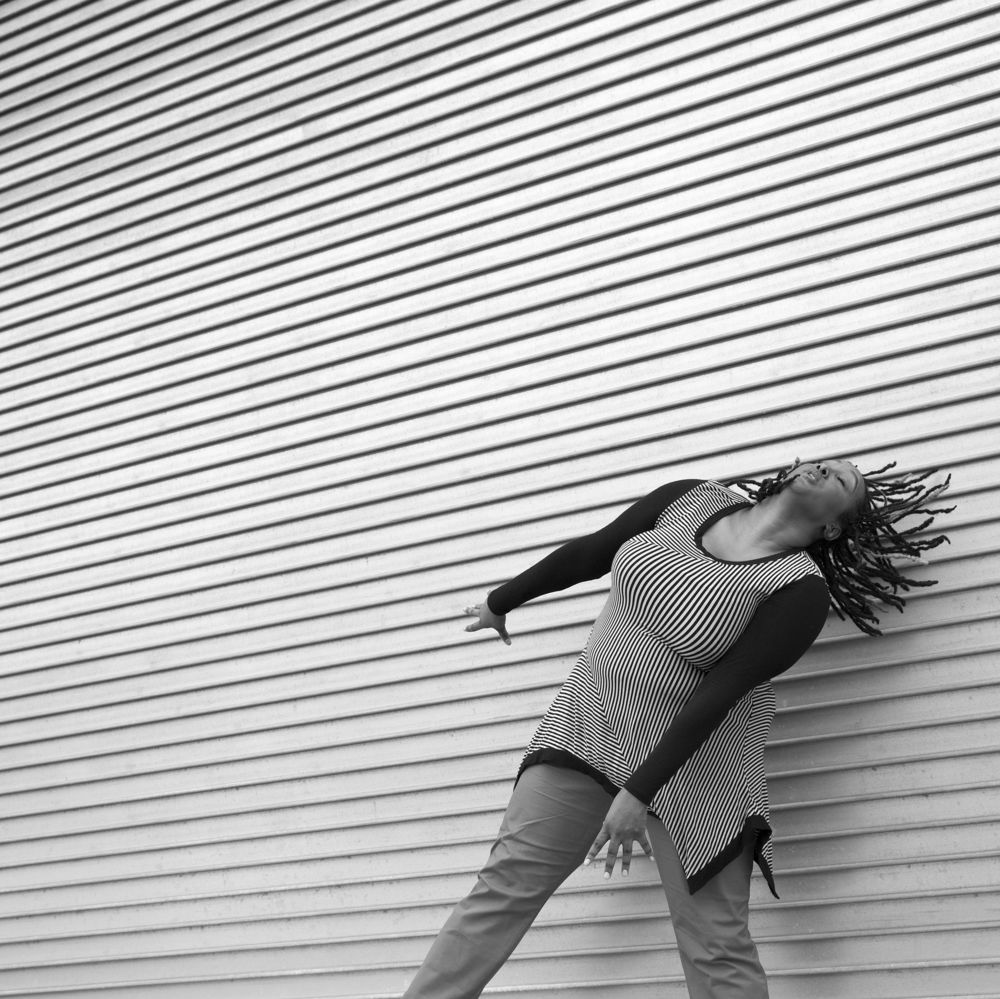 A black and white image of Kayla Hamilton, who is a milk chocolate-skinned Black woman. She is dancing in front of a textured wall that has horizontal layers. Her arms are energetically reaching down towards the diagonal as her head is tilting back with her locs moving back in that same direction. She is wearing a striped long sleeve shirt with pants.