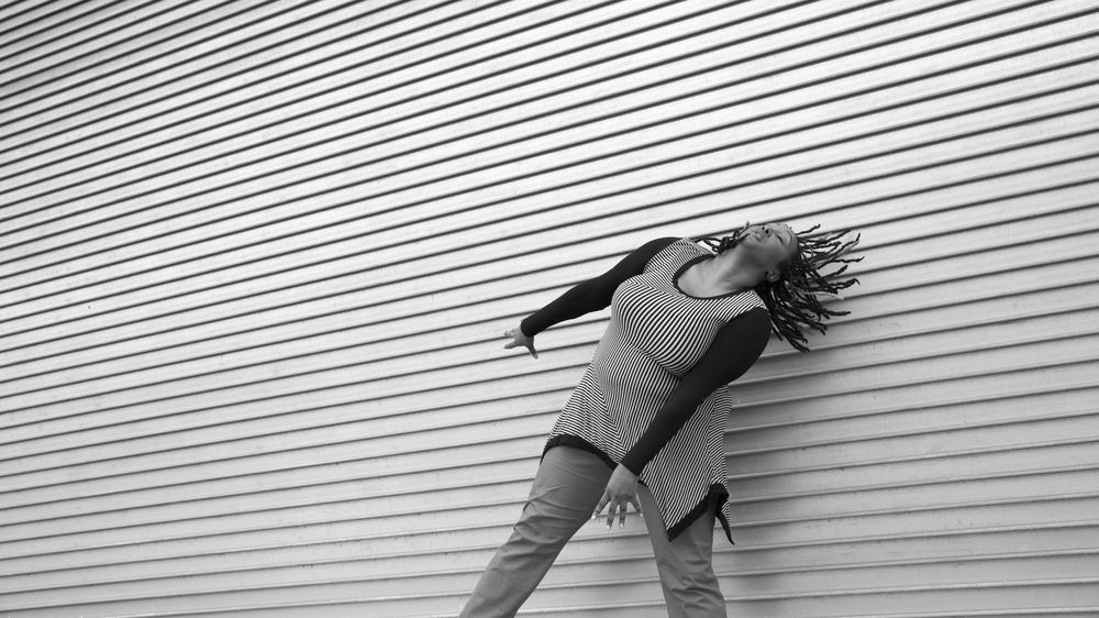 A black and white image of Kayla Hamilton, who is a milk chocolate-skinned Black woman. She is dancing in front of a textured wall that has horizontal layers. Her arms are energetically reaching down towards the diagonal as her head is tilting back with her locs moving back in that same direction. She is wearing a striped long sleeve shirt with pants.