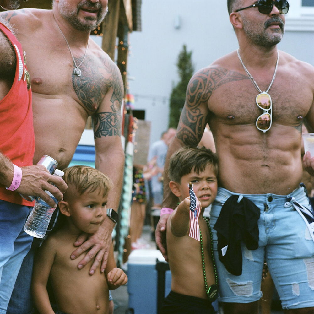 A square photograph of two muscular men standing outside; their torsos are tan and tattooed. Between the men are two small shirtless boys, around age five. The blonde boy on the left looks off to the right. The boy to the right brandishes a miniature American flag, aimed at the viewer, with one of his eyes closed in concentration.