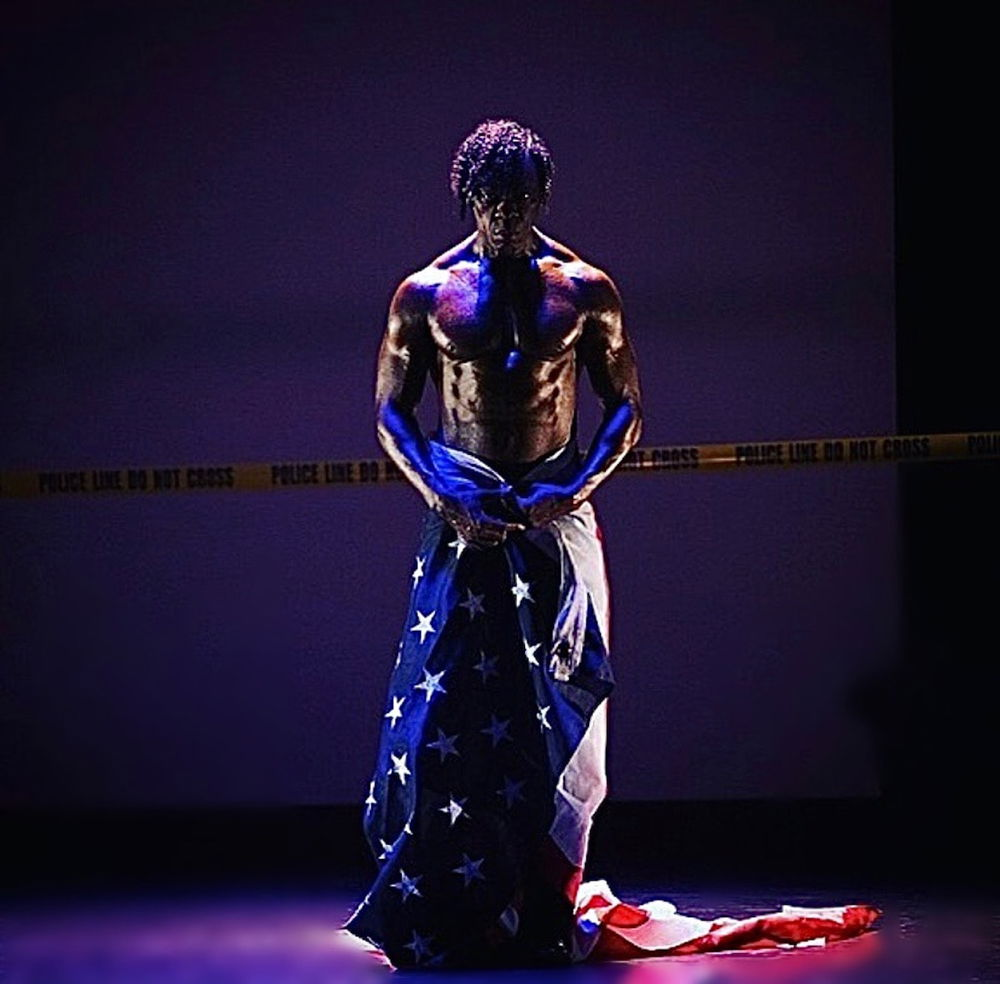A Black man stands under a spotlight, staring directly at the viewer. His gleaming torso is bare and his lower body is wrapped in an American flag. Behind him is a blank wall with a single strand of police tape running along the length of the wall.