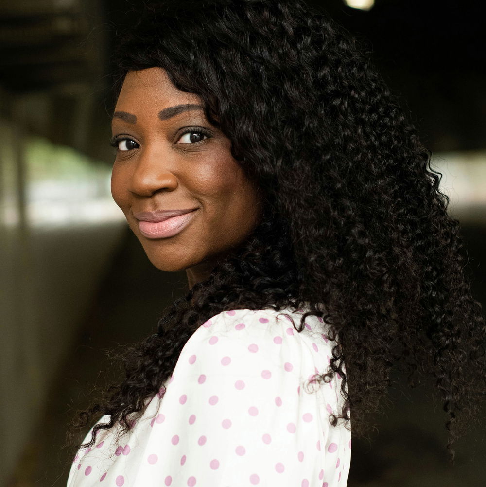 Natasha, a dark-skinned Black woman with Afro curly hair, wears an ivory and pink polka dot blouse and smiles.