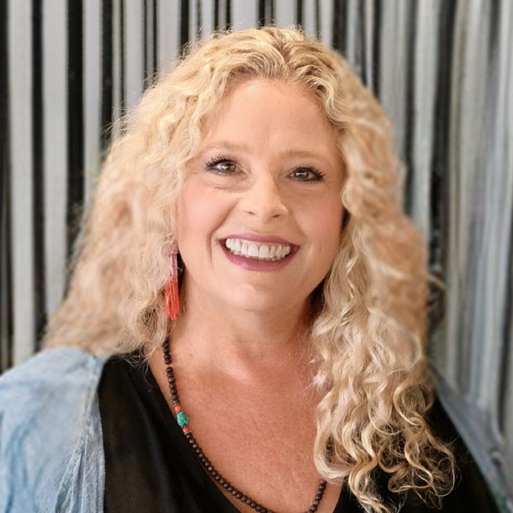 Beth, a woman with long curly blonde hair, faces forward and smiles directly at the camera. She is wearing a dark shirt, beaded necklace, and fringed earrings.