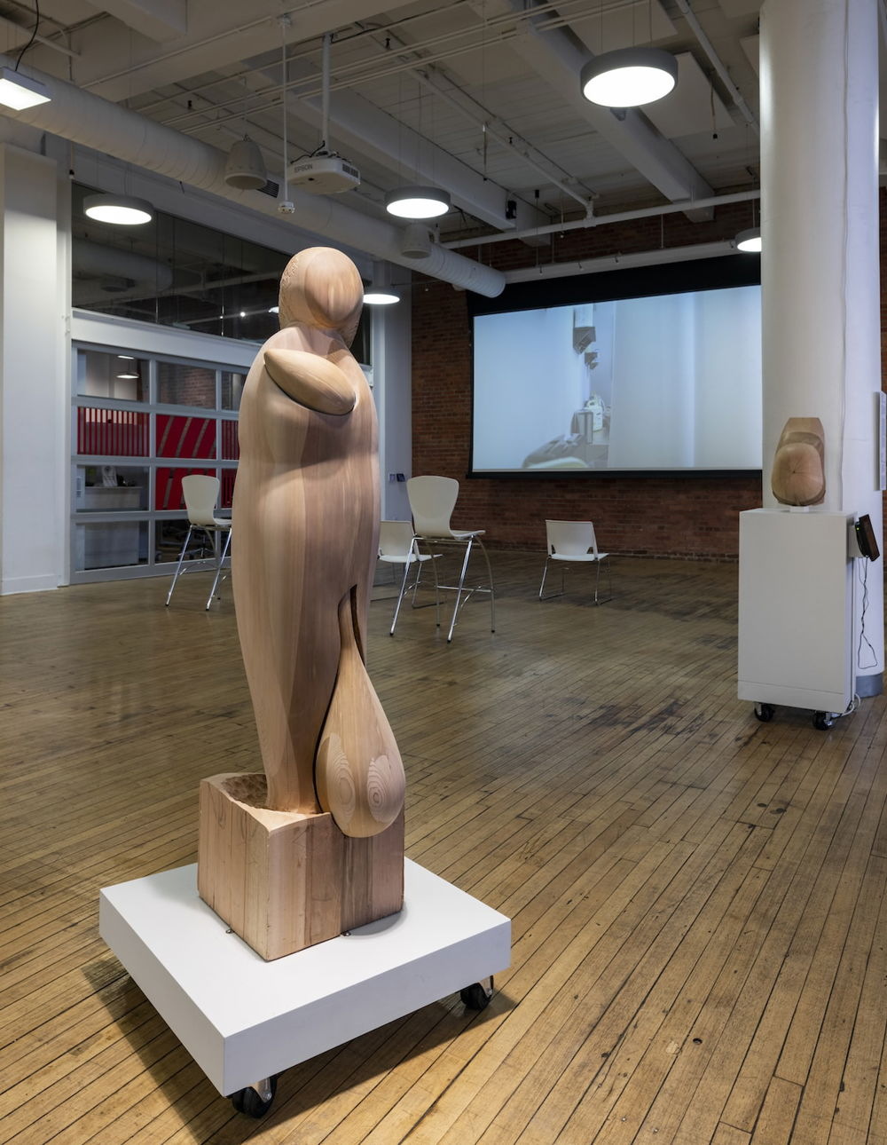 Photograph of a wooden sculpture in an exhibition space. The sculpture is in the shape of a person on a pedestal and has a smooth surface with additional pieces attached — one that resembles an infant in arms, and larger, rounded piece attached at the bottom that resembles a sack.