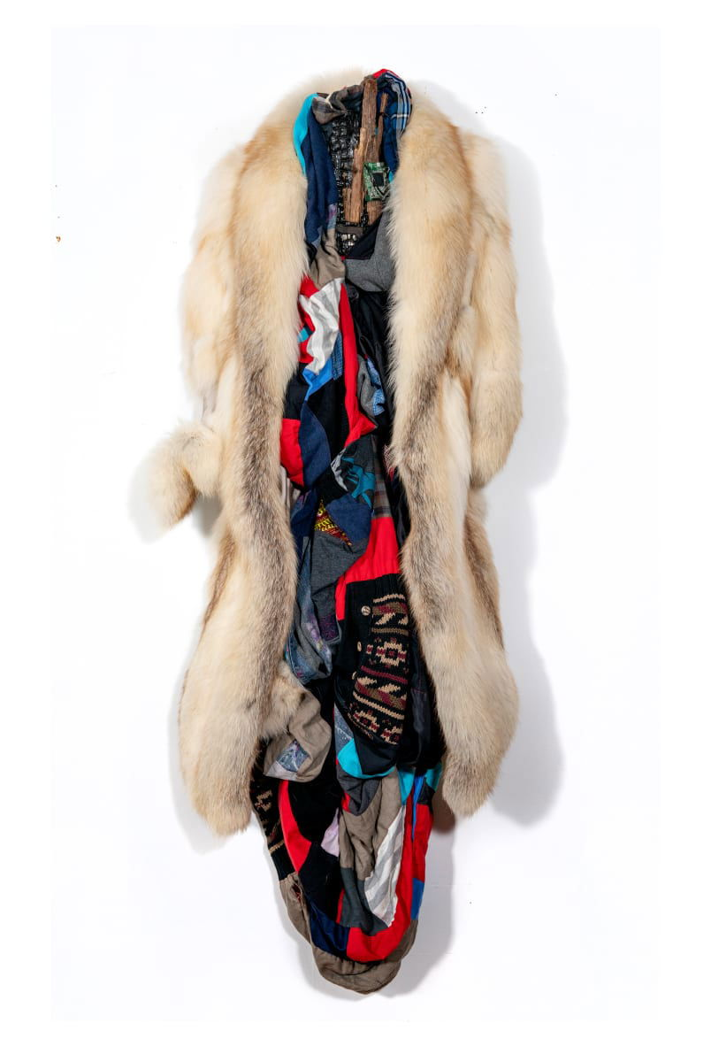 A mink fur coat is draped over a body of quilt. The red, blue and patterned quilt hangs within the confines of the coat and extends below the bottom of the coat’s length.