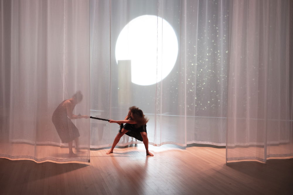 Two performers seem to play tug of war with a piece of black rope on a stage hung with shimmery, translucent curtains. The performer on the left is partly obscured by one of the curtains, as is a bright, circular light behind them, which looks like the moon.