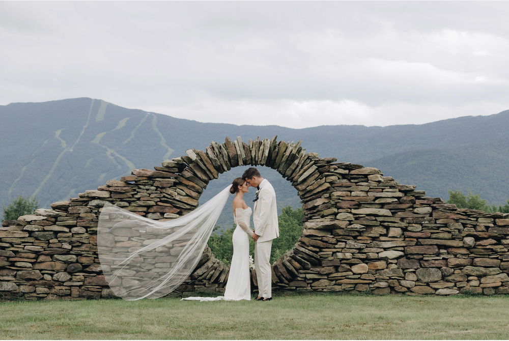 A bride and groom stand in front of a hand-built stone arch with a circular window built within. In the distance are tall mountains.