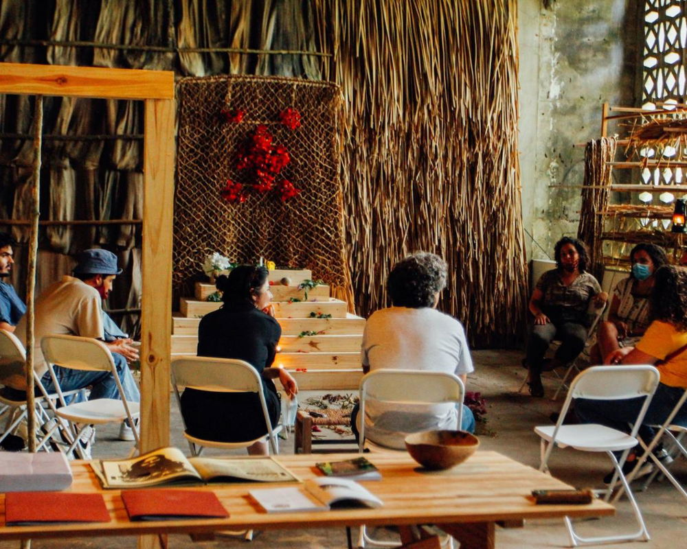 A photograph of a studio space with an installation made of natural materials strung onto the wall and a wooden altar, its steps scattered with flowers. A number of people sit in a row of folding chairs surrounding the altar space, engaged in quiet conversation.