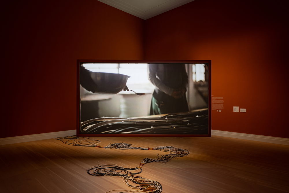 A framed TV monitor fills the corner of a windowless gallery space with terracotta red walls. The large, horizontal screen shows an image of a backlit hand holding a large silver mixing bowl and spoon hovering over a pile of wires. Outside of the monitor is the same bundle of wires that trail along the gallery floor.