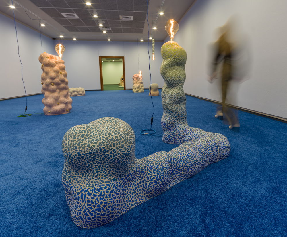 Large-scale, abstract ceramic sculptures are arranged in a room with blue carpet, each with a large lightbulb affixed to the top emitting a warm glow. In the foreground, an elongated sculpture stretches across the floor before jutting up toward the ceiling.