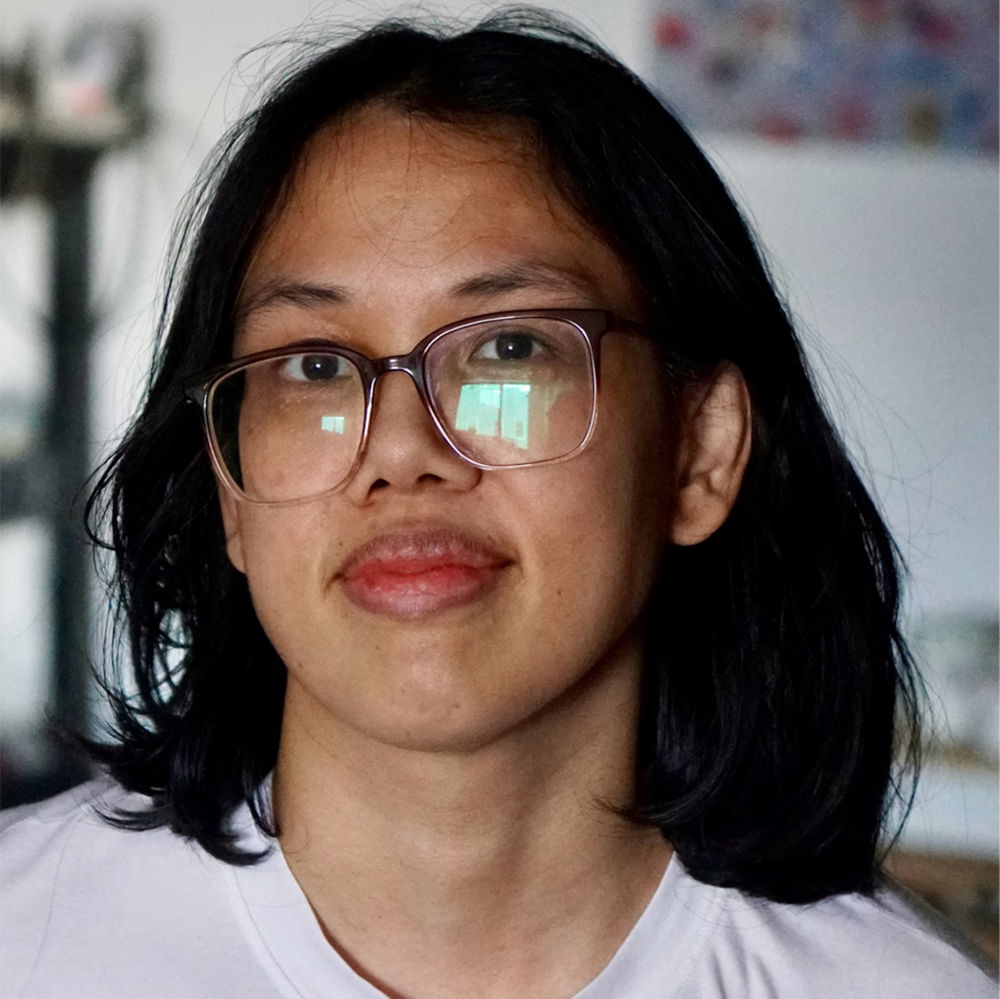 A tan-skinned person is pictured wearing gradient acrylic eyeglasses with clear lenses. Their shoulder-length black hair frames their face against a mostly white background.