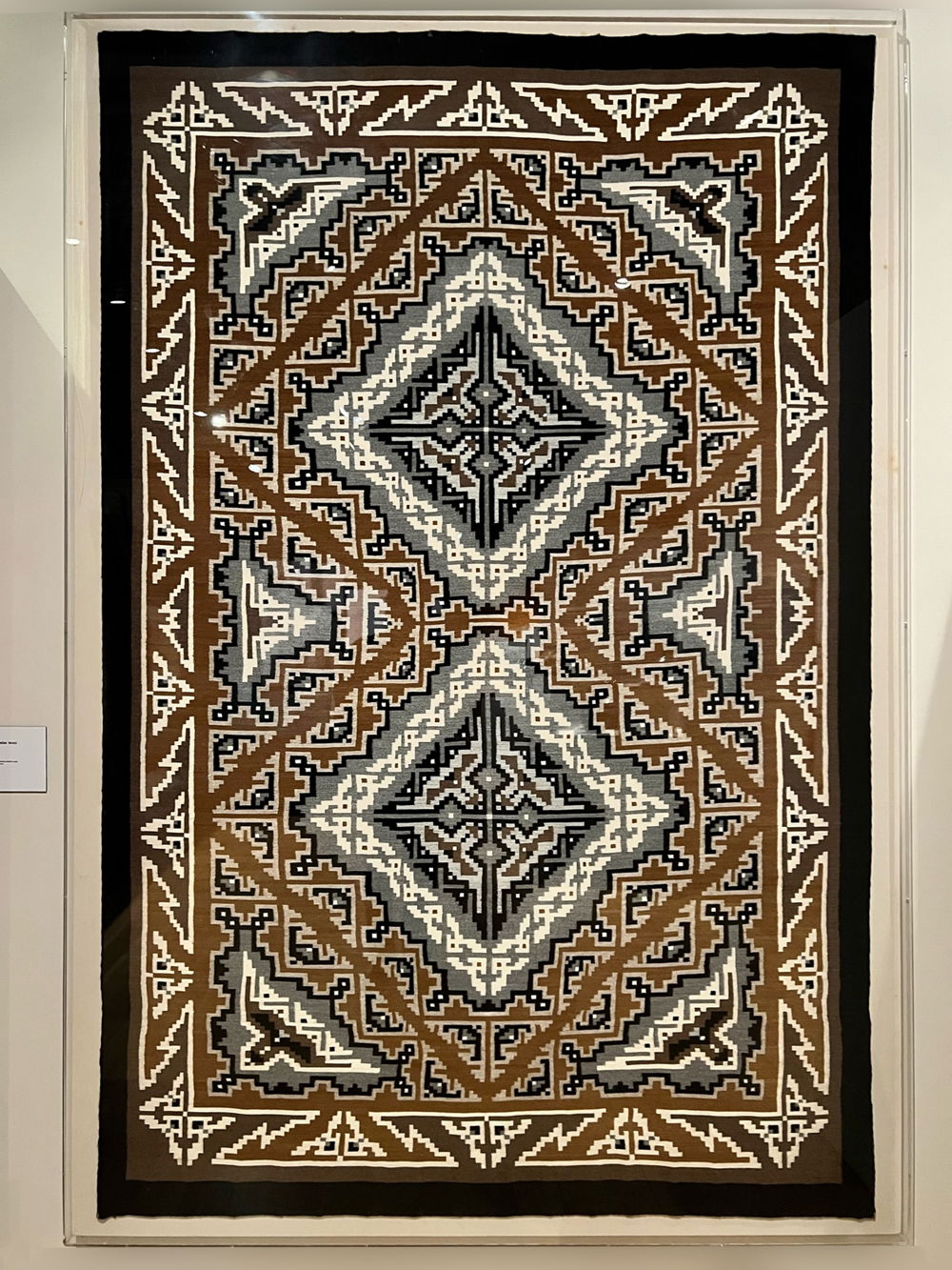 An intricately-patterned weaving in warm browns, whites, and grays with two central diamond shapes in the middle. Geometric patterns that resemble birds in flight point outward at each corner of the rectangle. Abstract patterns fill the space, drawing the eye around the artwork.