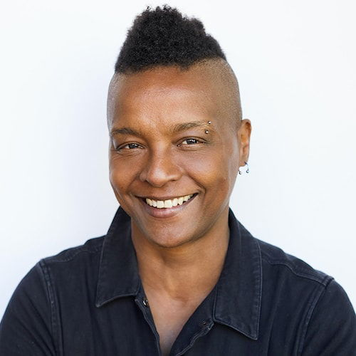 A headshot of a black, masculine-of-center queer person with a faux hawk style haircut. She is wearing short sleeved black coveralls with her arms folded. She is smiling.