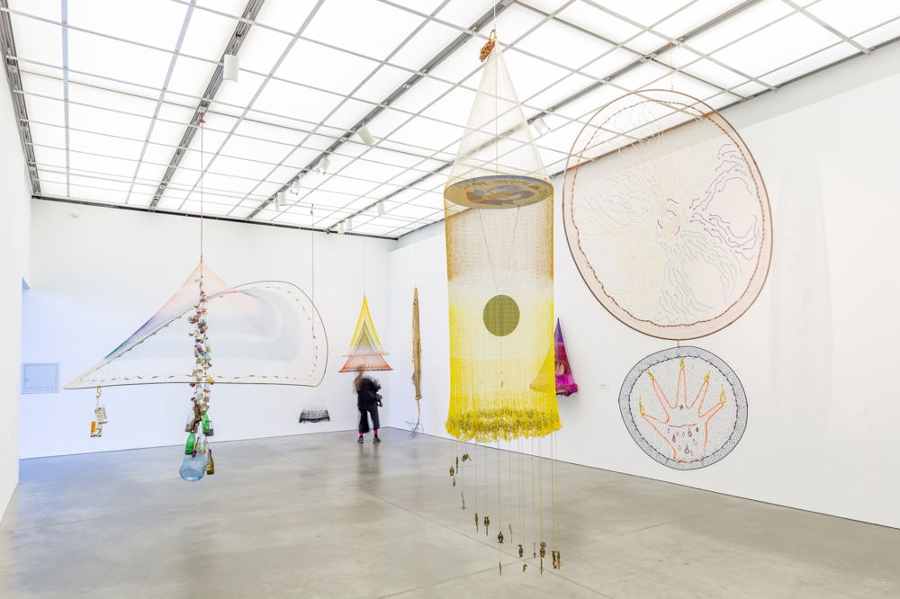 Photo of a white-walled gallery hung with brightly-colored mobile sculptures woven from various materials. The installation contrasts wide, airy structures against more solid pieces such as hanging medallions and a long, hanging sculpture composed of glass and plastic bottles.
