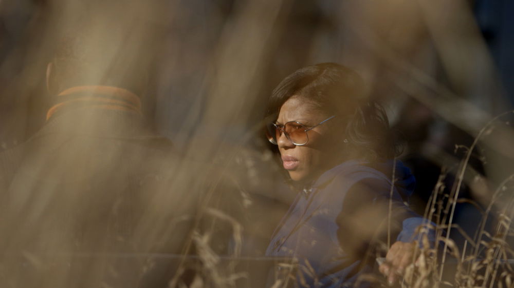 A woman in sunglasses stands in tall grass, her face partially turned to the camera. Out-of-focus blades of grass in the foreground partially obscure another person to her left.