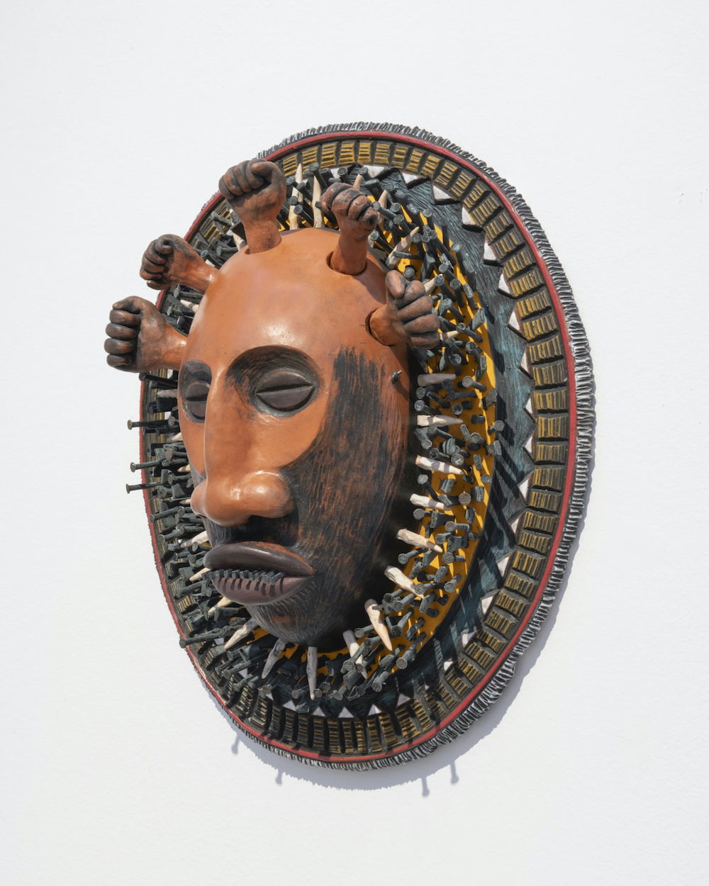 A mixed media mask displayed on a circular wall platform. The mask depicts a stylized face and is a terra cotta color with black accents around the lower half of the face and closed eyes. Five miniature fists crown the top of the head like small tufts of hair. The circular platform is ornately patterned with geometric shaped pieces of mixed media materials such as ceramic and metal pieces.
