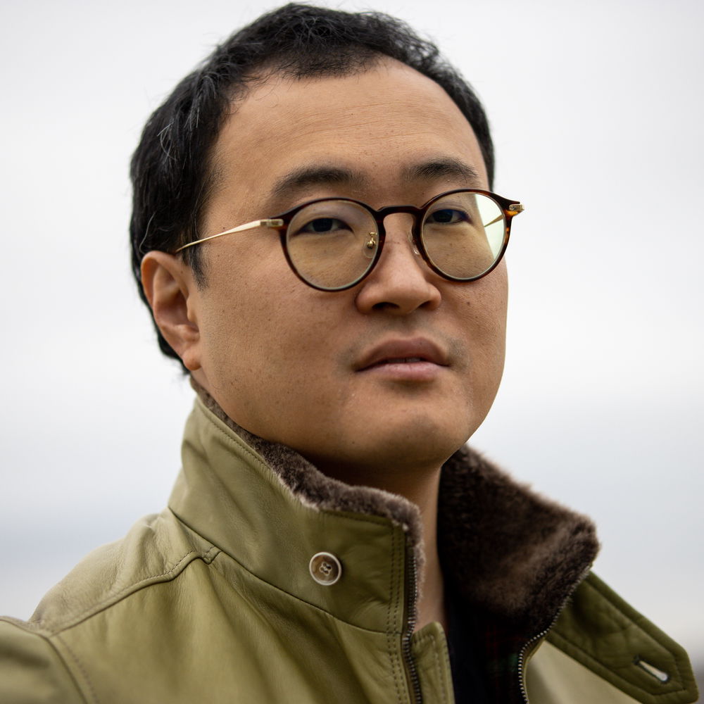 An Asian man with short black hair in his forties poses outside on a gray, cloudy day. He is wearing tortoiseshell horn-rimmed glasses and a green leather jacket. 