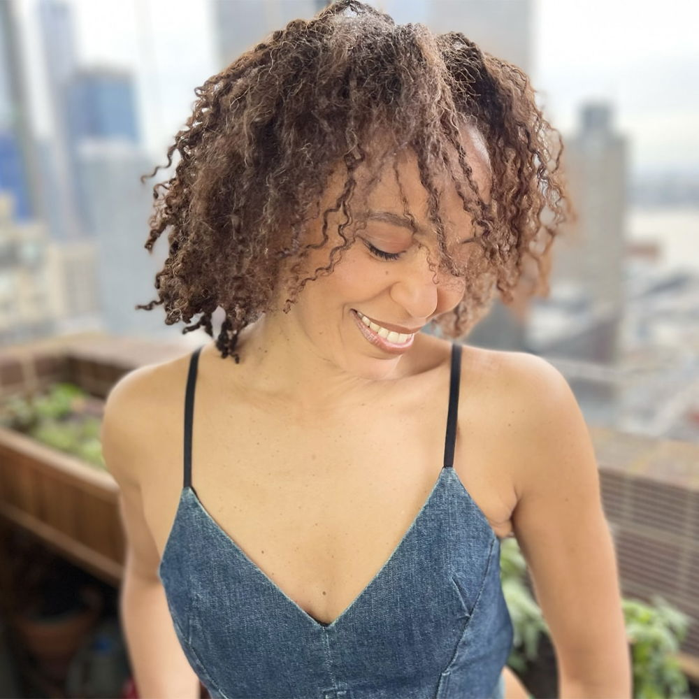 a black woman with curly hair wearing a denim tank top smiles while looking down on a balcony overlooking the skyline of Mannahatta.
