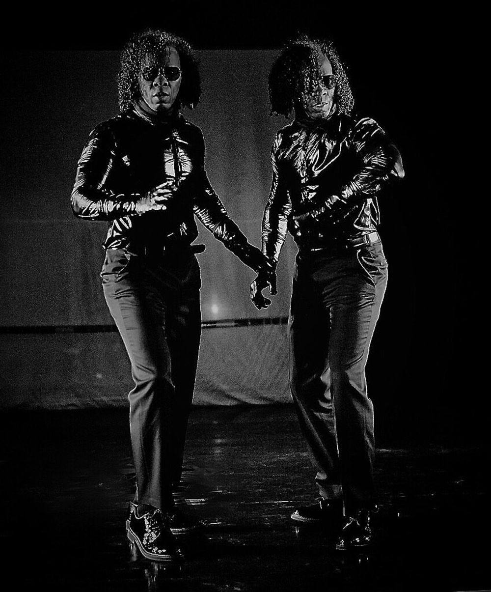 A black-and-white image of two figures standing stiffly in a dark interior. Both figures have curly mid-length hair and are dressed identically in dark pants, shiny jackets, and dark glasses. The figures look in different directions but are in mid-motion as if startled.