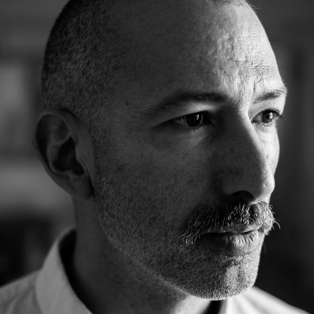 A black-and-white portrait of a middle-aged Syrian-Peruvian man with buzzed salt-and-pepper hair and mustache. He is wearing an Oxford button-down shirt and gazes out beyond the frame.