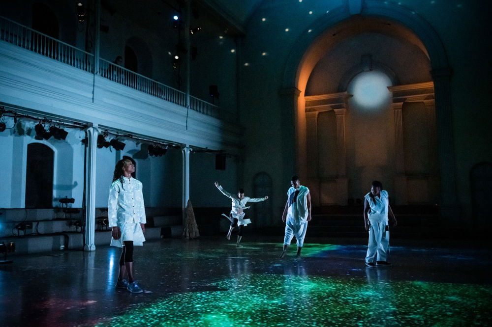 Four dancers wearing white glow in darkened space, which is illuminated by galactic projections on the floor. One dancer jumps with her hands and arms stretched wide.