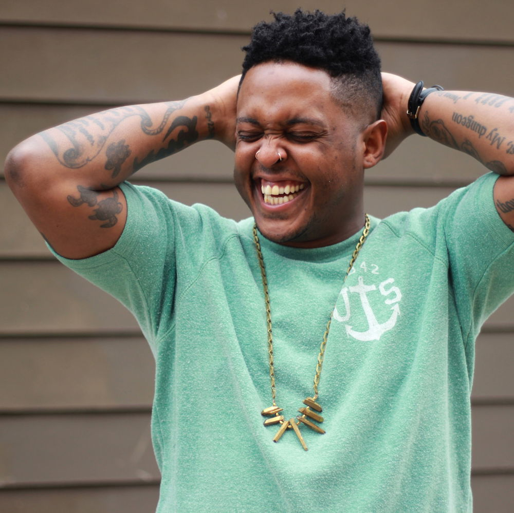 Danez, a young Black person with a short high top fade, smiling with all their teeth. They are wearing a green shirt, a multi-colored tank top, a gold necklace, and a wristwatch.