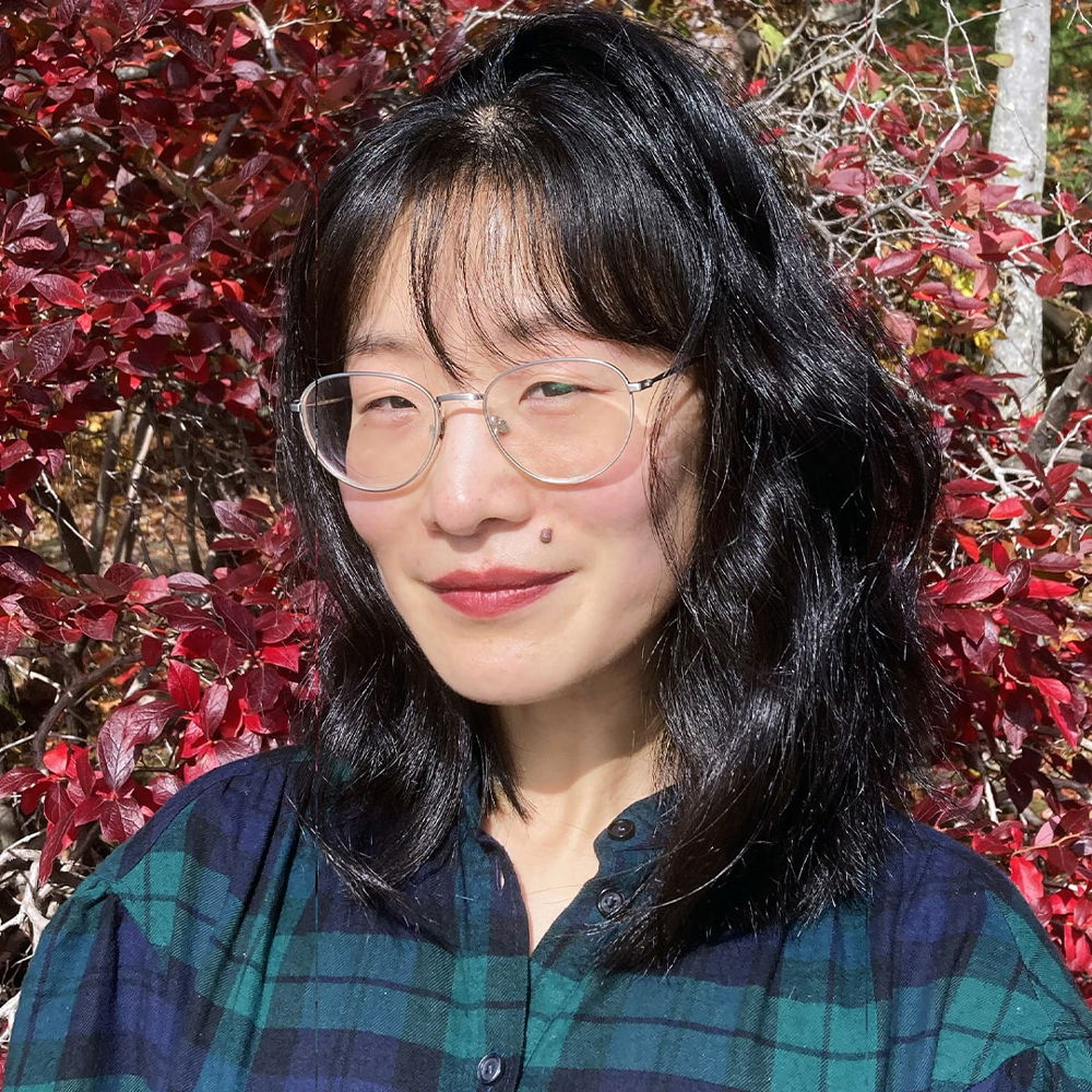 A Chinese woman with dark, shoulder-length hair and silver-rimmed glasses stands in front of the red leaves of a sourwood tree. She looks into the camera smiling.