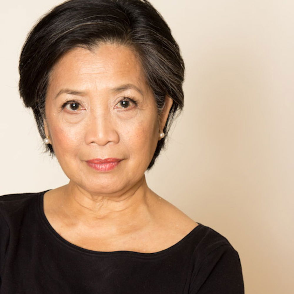 Mia, an Asian American with short black hair, is wearing a black scoop-necked shirt against a light background. She stands with her arms crossed in front of her.