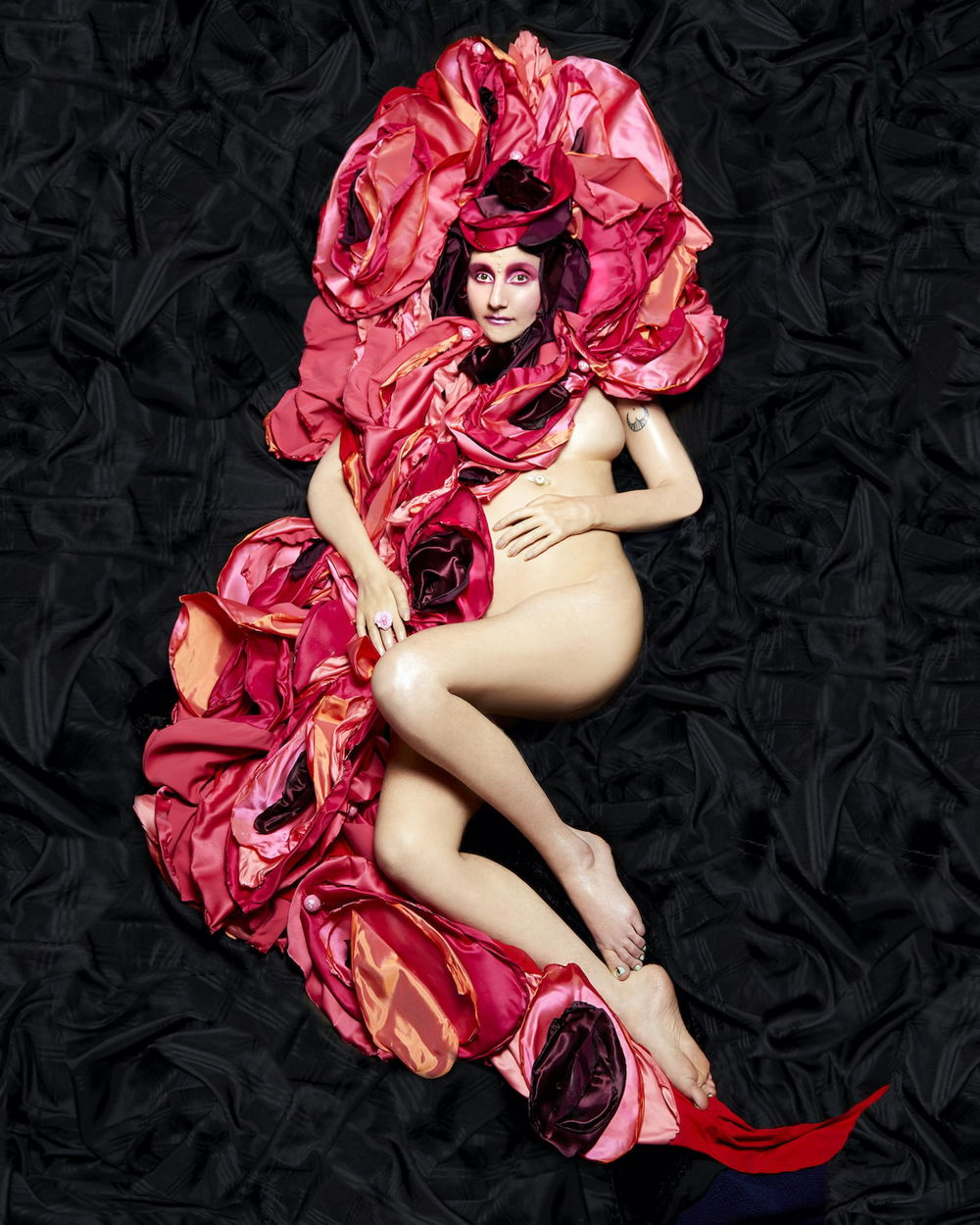 A nude White woman luxuriates on a background of black fabric. The left side of her torso is engulfed in the deep folds of pink fabric that circle around her head and is tucked under her bent knees.