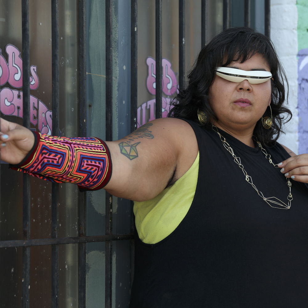 Mother Cyborg, a young Latin woman with shoulder-length black hair, poses in front of a colorful street wall in cyborg-like white shades, a black sleeveless T-shirt, and neon colored printed arm bands.