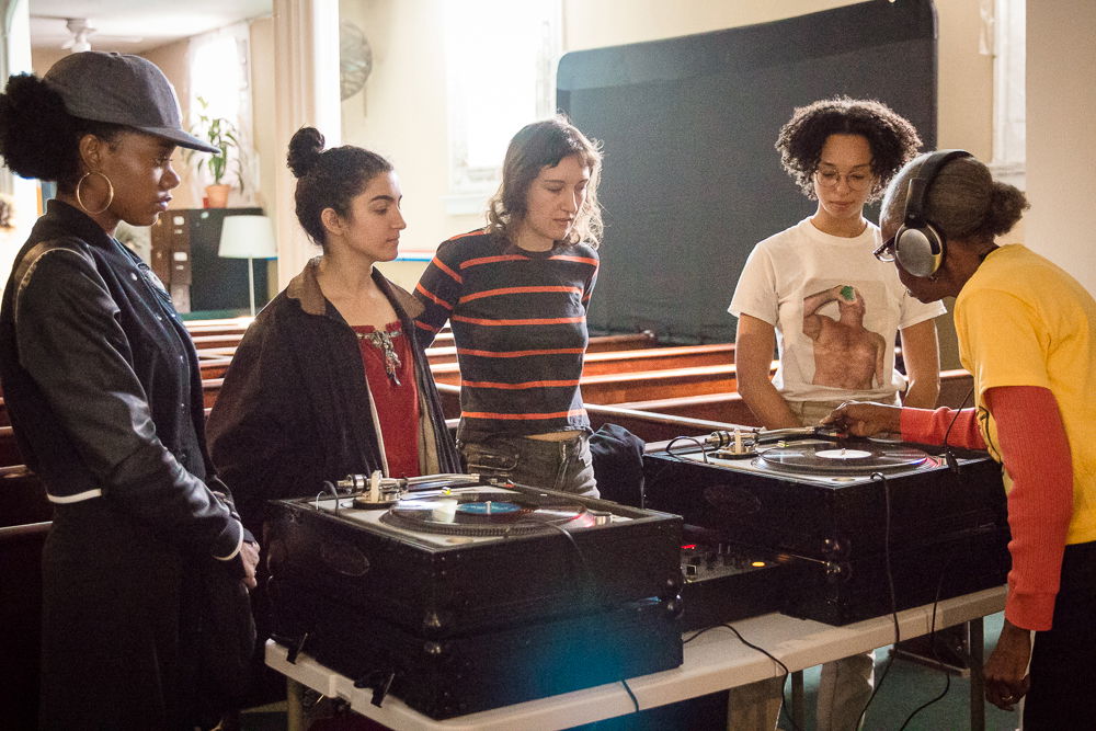 Four young women watch an elder Black woman DJing with vinyl records. The DJ has headphones on and drops the needle onto a record.