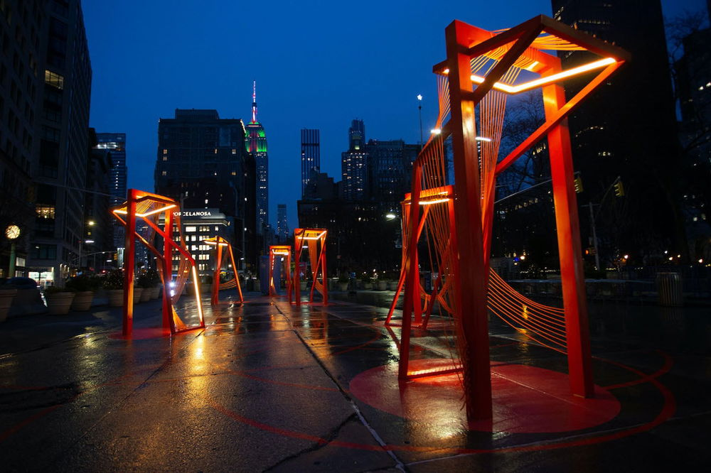 Photo of an outdoor art installation at night. Six bright red architectural sculptures are constructed on the cement of a public plaza, illuminated by lights. The frames of each structure are strung with ropes, to create an inviting space.