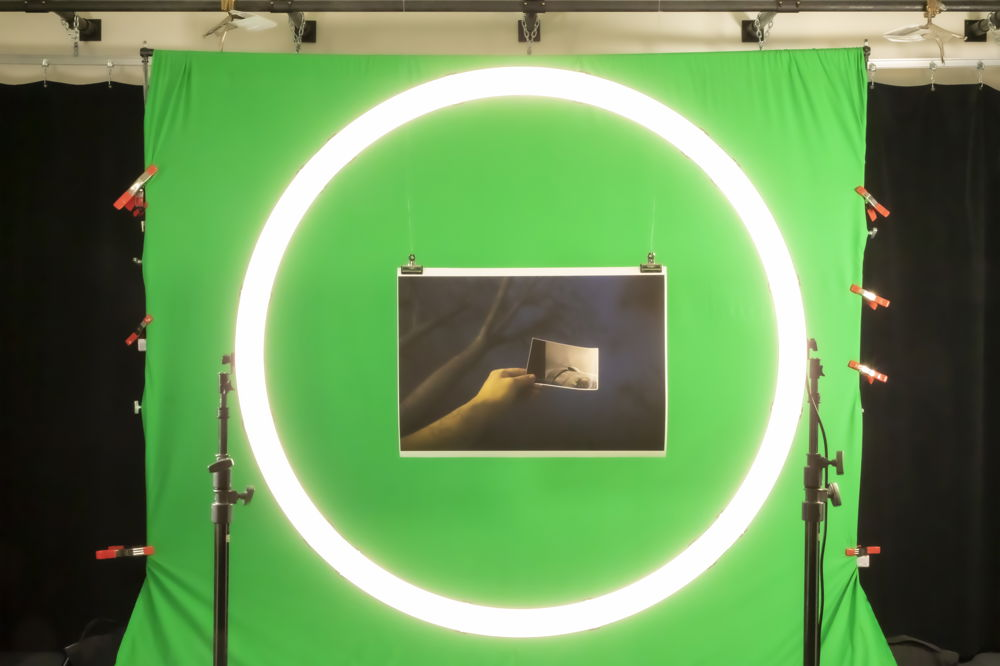 An elaborate documentation set up where a work of art is placed in front of a green screen and photographed through a ring light. The artwork itself is a photograph of a dark forest with a hand reaching upward toward the trees holding an even smaller photo.