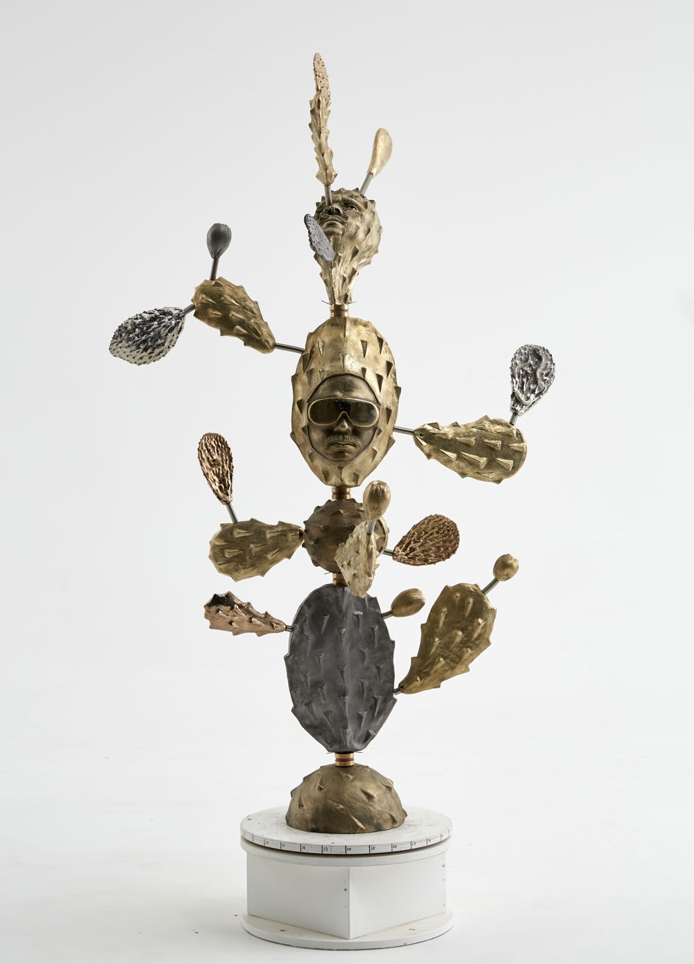 A totem-like brass sculpture with a combination of cast iron, gold and rose-gold plating prickly cactus paddles. The center figure is a cactus with a portrait of the artist wearing futuristic polished glasses.