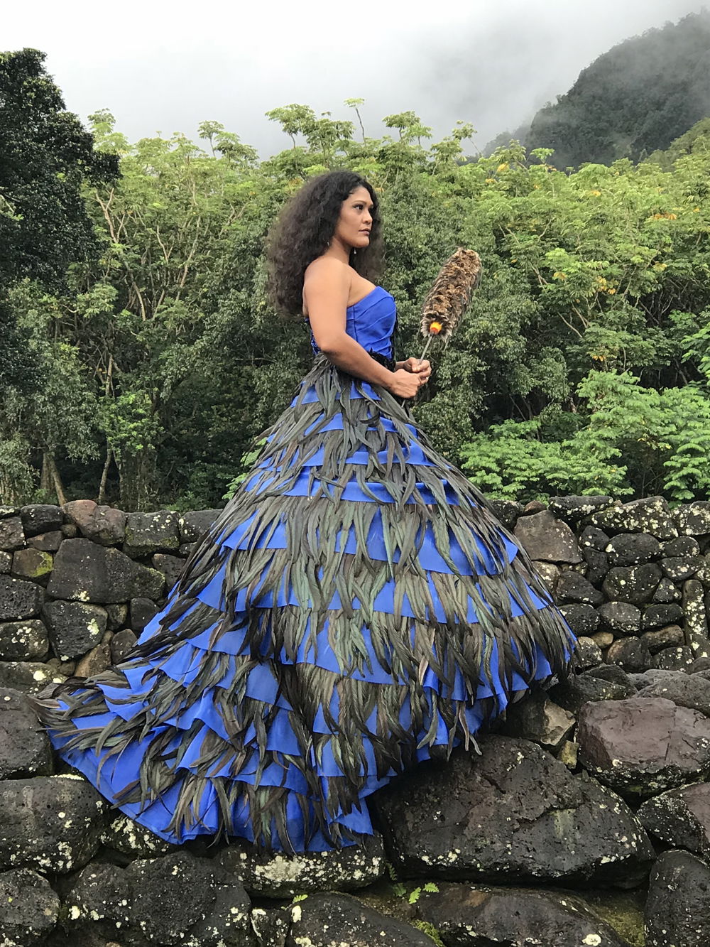 Standing on black rocks and backdropped by mountainous trees, a model with tan skin and long curly black hair models a royal blue ball gown circled with rows of long greyish green feathers.