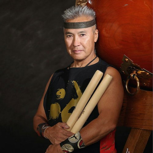 Portrait of a man standing in front of an odaiko. The drum fills the right half of the image, and the man rests his head and back on the instrument. His distinguished short, grey hair is pushed upward by a brown headband. His muscular arms rest at his midriff and hold two wooden mallets.