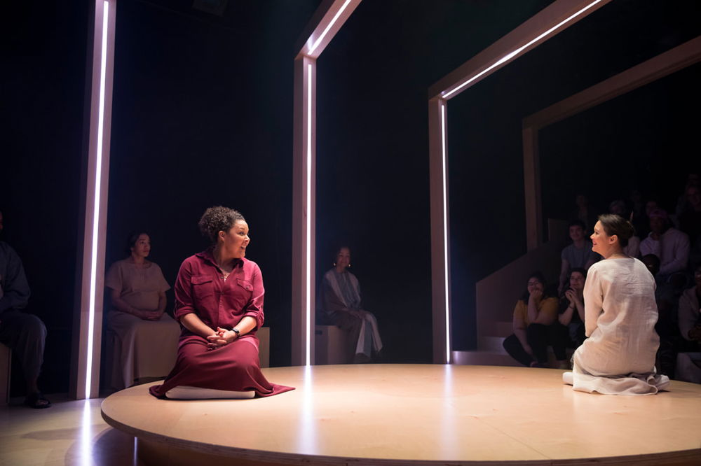 Two actresses sit on a circular stage, across from one another. Above the stage are rows of rectangular structures that emit soft pink light.