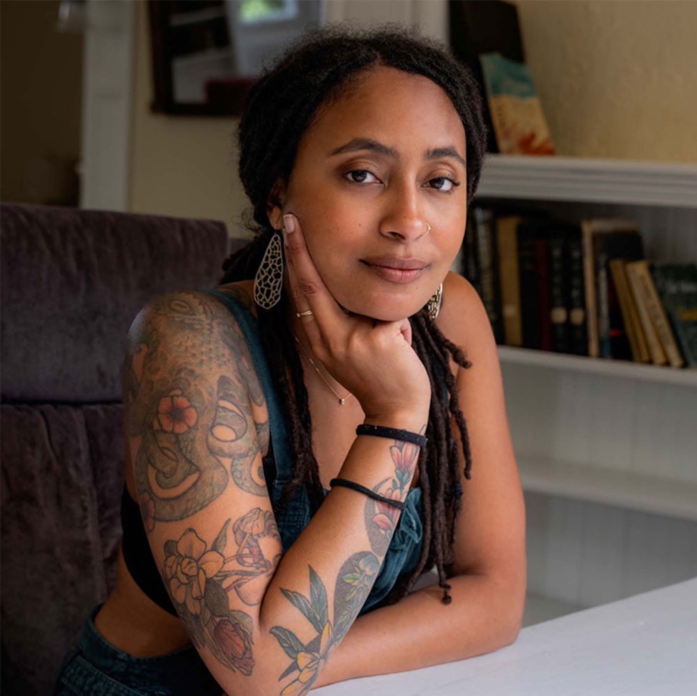 A Black woman with long locs pulled back from her face and a light-brown complexion sits at a desk in front of a bookshelf backdrop. She wears a pair of gold mesh-work earrings and green overalls with a black crop top underneath, baring her sleeve of colorful tattoos.