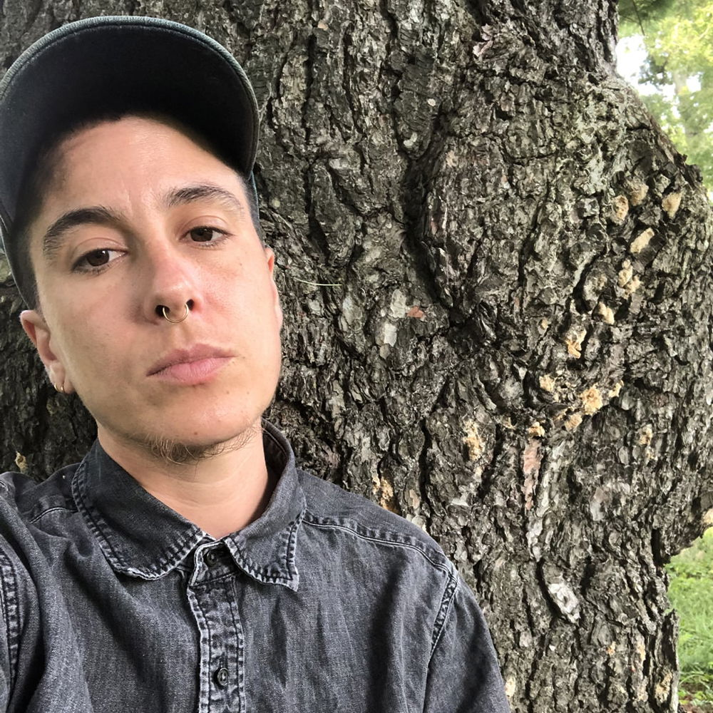 A sun-skinned, mixy transmasculine person rests against a tree with a ball cap on looking directly into the camera. They have scraggly chin beard, gold septum piercing, and they wear a denim gray button-down shirt.