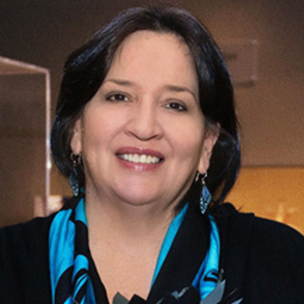 A photo of a woman with dark brown hair and a happy smile. She wears a blue scarf, a black shirt, and dangling earrings that resemble butterfly wings.