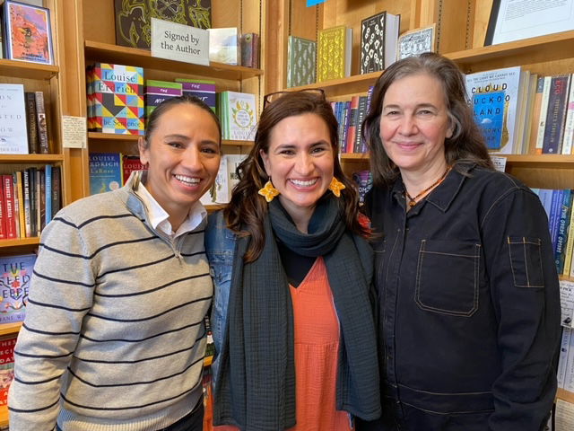 Louise stands on the right with two women on her left who smile broadly at the camera. They are framed in front of shelves lined with many books.