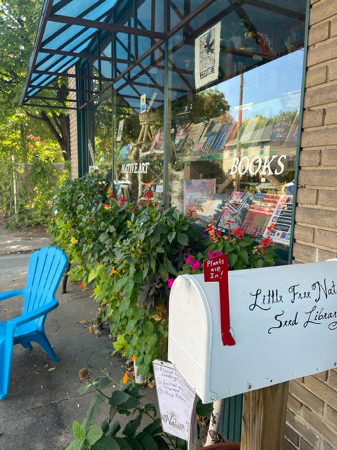 Photo of the outside of Birchbark Books store. Many books are on display behind the glass windows. Green plants and flowers are lined up under the windows, and a blue chair sits out front. A white mailbox with the words “Little Free Native Seed Library” hand-painted on the side stands up in the foreground.
