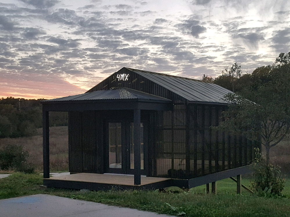 A photograph of a one-room structure with a bit of woods and a sunset visible behind it. It is made of corrugated plastic and black beams with a small porch at the front. At the top is a white neon sign that reads “4mx."