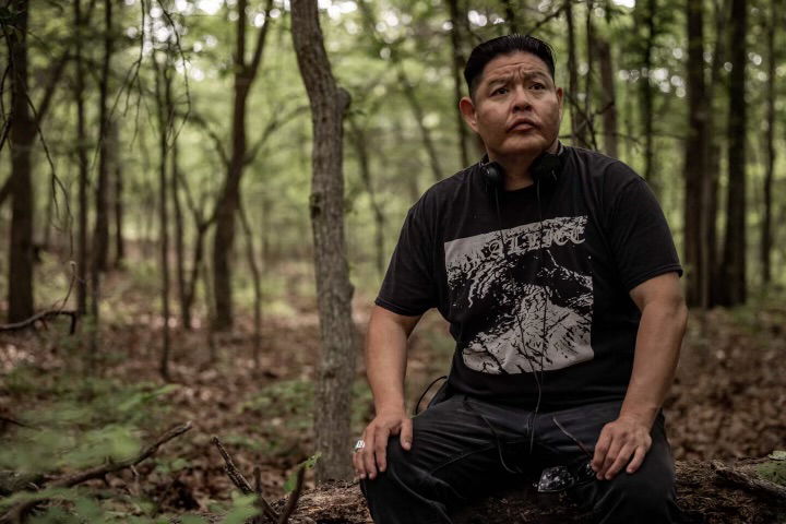 The man in the image appears to be contemplating or observing his surroundings in a dense, green forest setting. He has a serious, pensive expression on his face as he sits on a fallen tree trunk or rock. The natural environment with its towering trees and undergrowth creates a contemplative atmosphere.