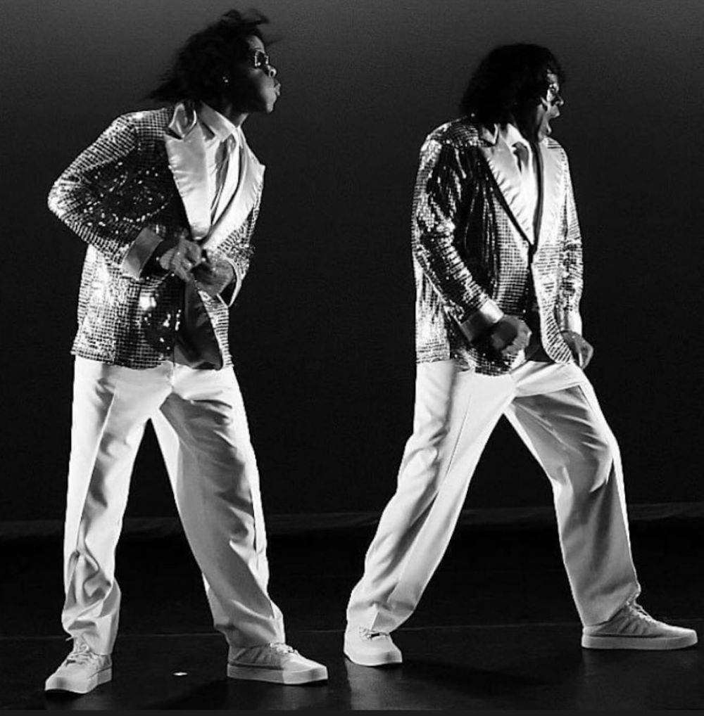 A black-and-white image of two figures on a stage. Both figures are dressed smartly in white pants and sequined jackets. Both are turned to the right, faces only seen in profile.