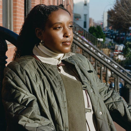 Portrait of an Ethiopian-American woman sitting on a brownstone stoop on a sunny Brooklyn day. She is looking away from the camera and wears a green puffy jacket with her hair in a ponytail.