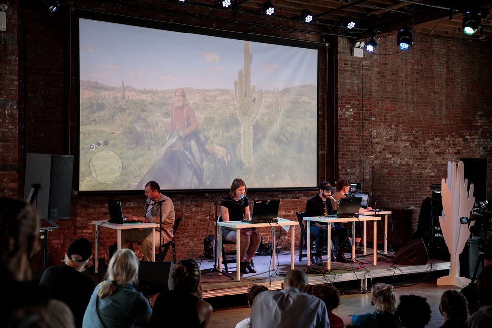 In a spacious brick room, four people sit at individual desks with laptops on a shallow stage, reciting out towards an audience on all three sides of the stage. Behind the performers is a large screen with a projection of a video game showing a man on a horse in a desert scene.